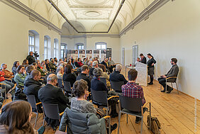 Mehrere Dutzend Personen (Publikum) und Vortragende bei der Eröffnung der Ausstellung. Das Publikum sitzt in einem Halbkreis vor den Vortragenden, im Hintergrund sind die Rollup-Banner der Ausstellung und Fenster sowie darüber die Stuckverzierung des Raumes zu sehen.
