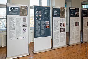 Mehrere blau-weiße Rollup-Banner der Ausstellung, die auf einem braunen Holzfußboden stehen. Im Hintergrund sind Fenster zu sehen. Auf den Bannern sind Namen von Personen, der Titel der Ausstellung, Fotos, eine Landkarte und Texte abgebildet.