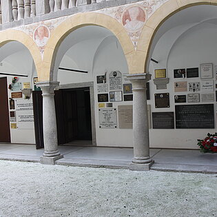 Auf dem Bild ist der Arkadengang in der Gedenkstätte. Man sieht Säulen, bemalte Rundbögen und Gedenktafeln sowie Blumengestecke.