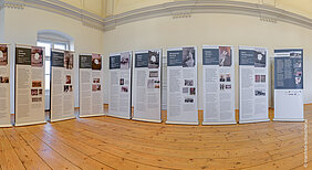 Mehrere blau-weiße Rollup-Banner der Ausstellung, die auf einem braunen Holzfußboden stehen. Im Hintergrund ist ein Fenster und über den Bannern eine Stuckverzierung an der Wand zu sehen. Auf den Bannern sind Namen von Personen, Fotos und Texte abgebildet.